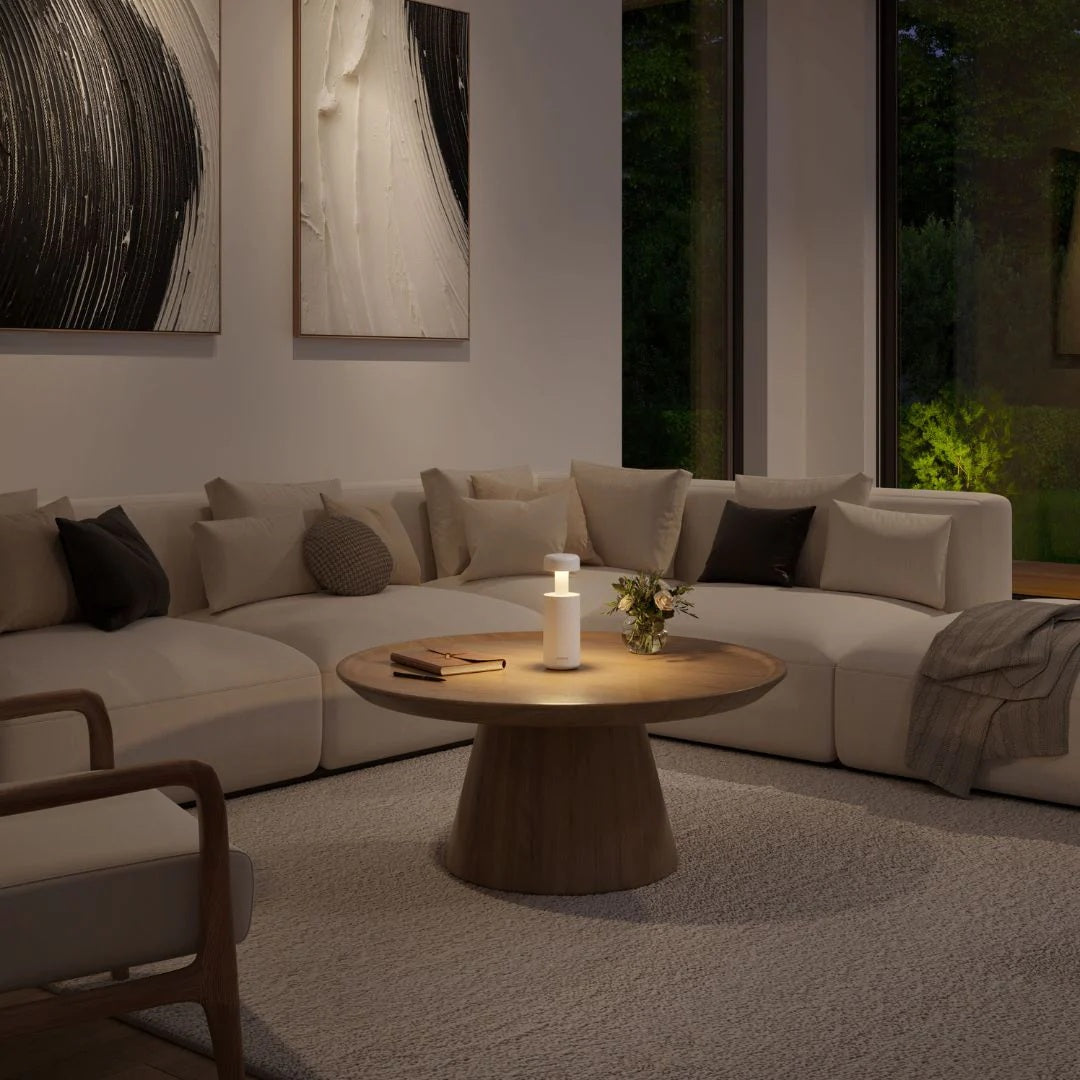  A modern Sompex Lubao table lamp with a warm glow sits on a round wooden coffee table in a contemporary living room, with a large beige sectional sofa and abstract wall art.